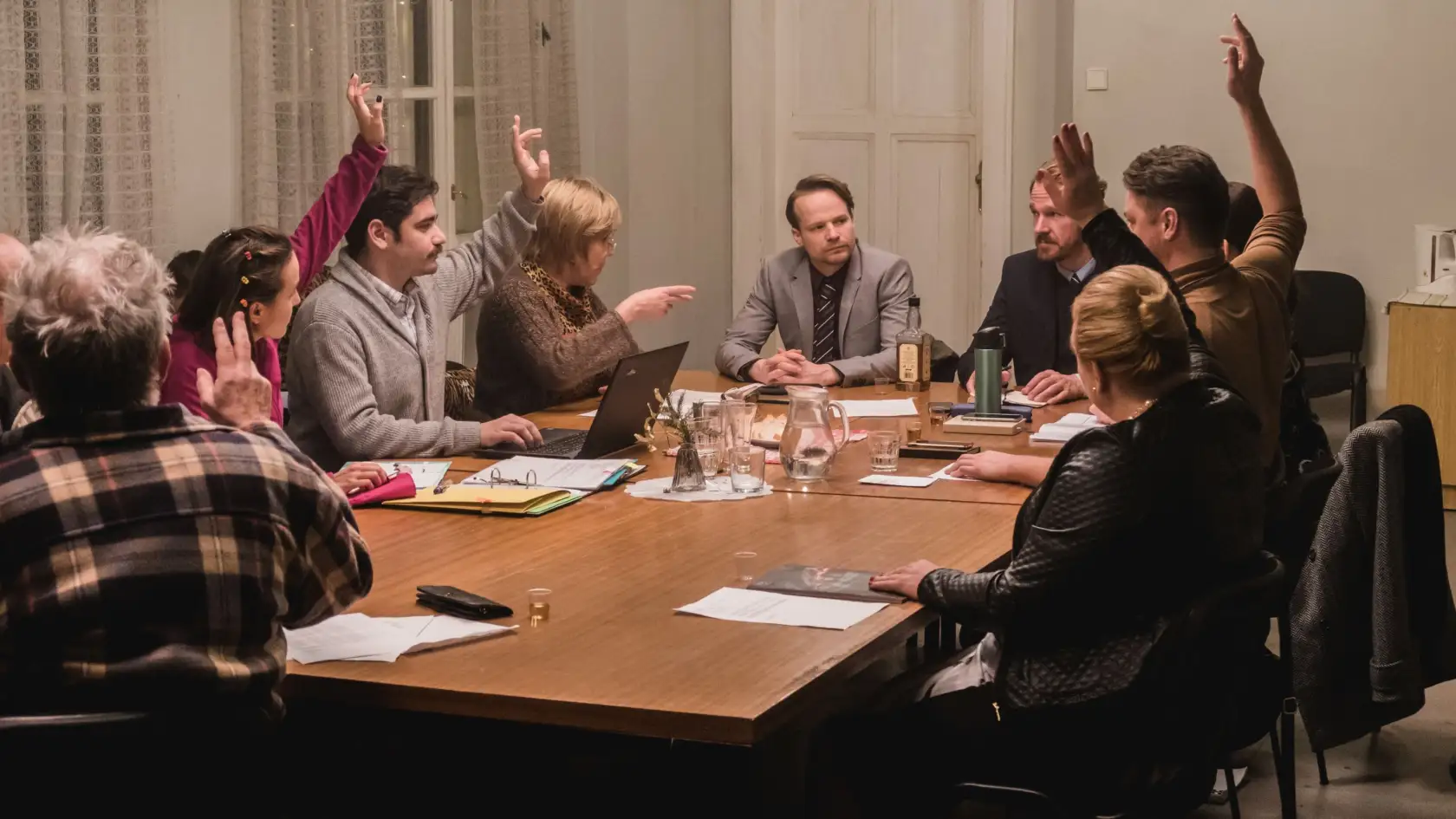 A group of people sit around a meeting table, some raising their hands to vote.